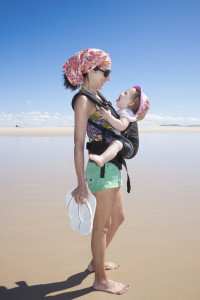 mother with headscarf green shorts carrying her one year baby with pink hat in front rucksack walking at a beach